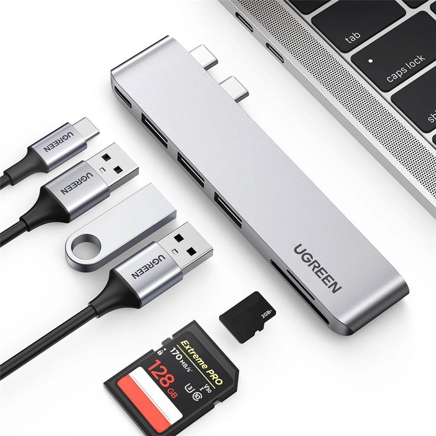 UGREEN 60560 dual USB-C hub in silver clipped to the side of a MacBook, with USB drives and SD cards beside it